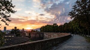 Veduta delle storiche mura di Lucca al tramonto, con il cielo che mostra un mix di colori sereni e nuvole suggestive. (Immagine generata con IA)